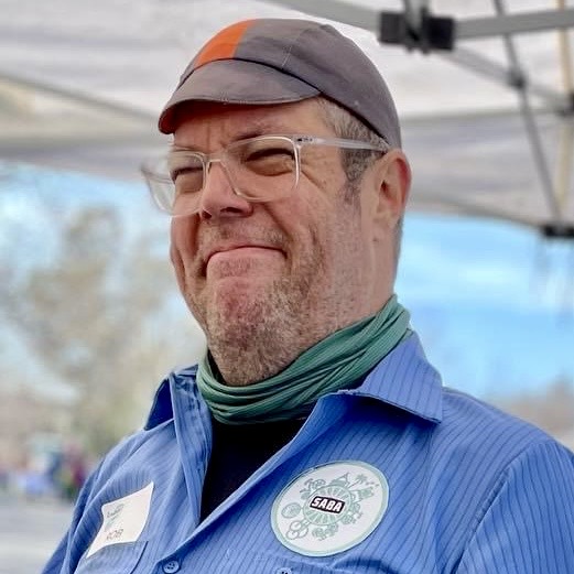 Headshot of a white man with a green cap and blue shirt and glasses.