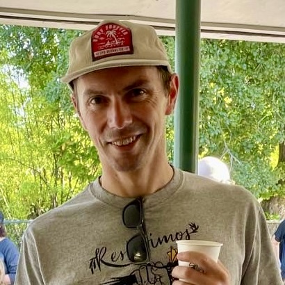 A white man wearing a beige cap and shirt holds a cup.