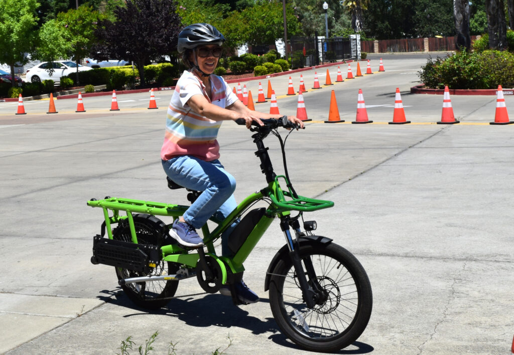 A person wearing a helmet and sunglasses sits on a green e-bike with an extended rack in a parking lot with orange cones.