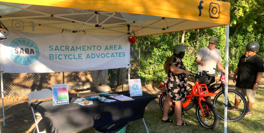 A booth set up outside with a banner that says, "Sacramento Area Bicycle Advocates." A woman holds on to an e-bike while two men look at it.