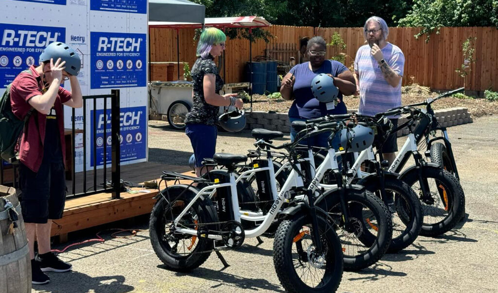 Four e-bikes lined up in a parking lot. Four people stand nearby, two wearing bike helmets.