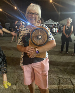 A white woman with with hair, glasses, pink pants, and a flowered shirt holds a board with a small bike wheel mounted on it.