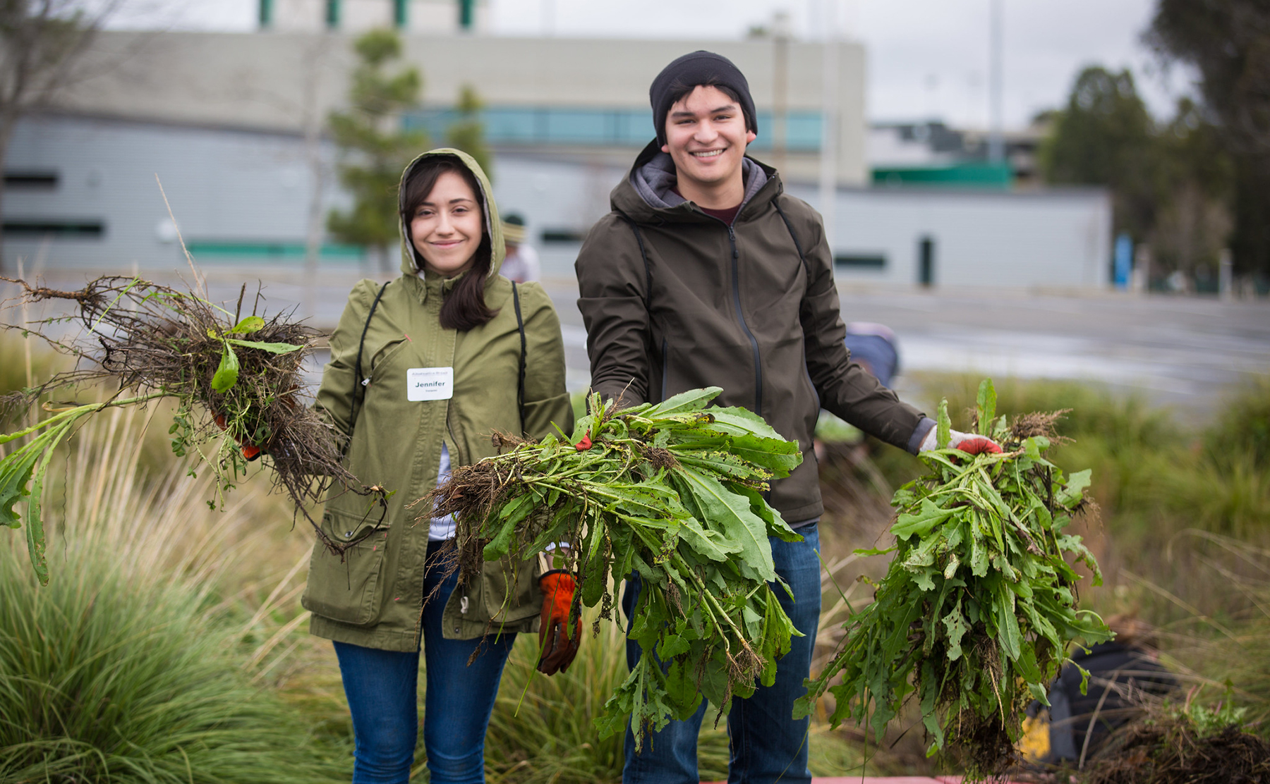 A young woman and a young man stand in a garden holding vegetation.