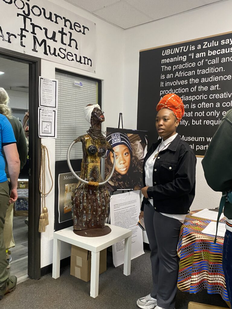 A young Black woman wearing a red scarf on her head stands by a sculpture at the entrance to the museum.