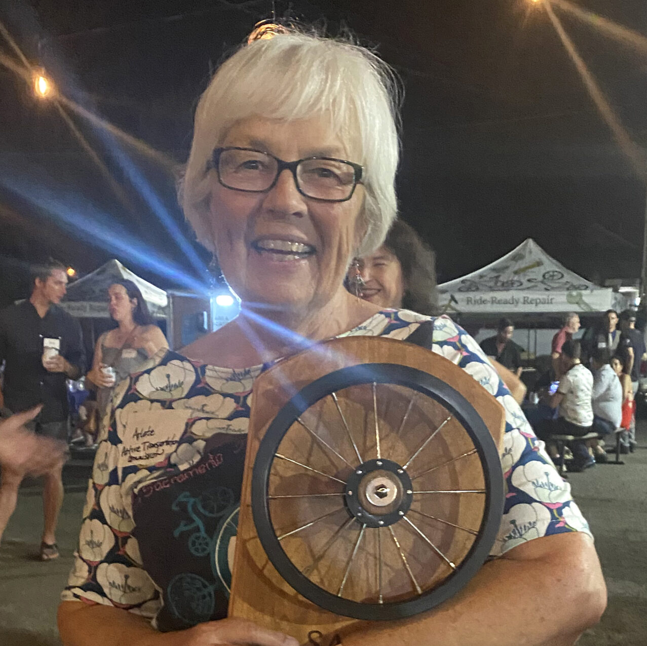 A woman with white hair and glasses smiles as she holds an award with a small bike wheel attached to it.