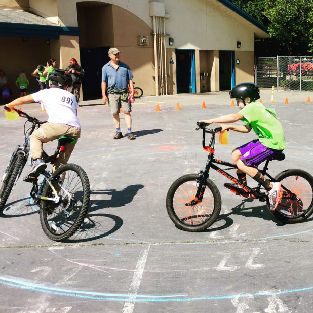 Two kids ride bikes in a parking lot while an adult looks on.