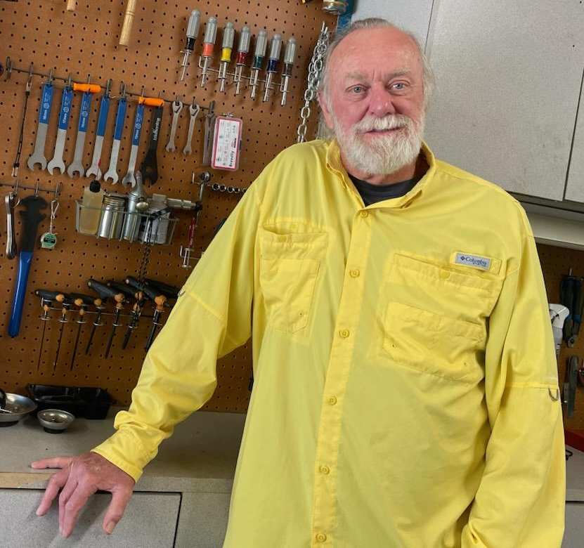 An older white man with a white beard and yellow shirt stands in front of a wall of tools.