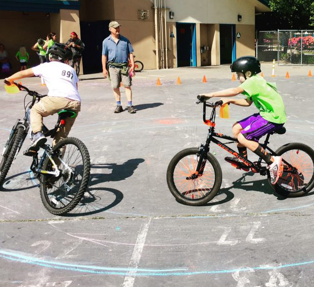 Two kids on bikes ride in a circle while a man watches.