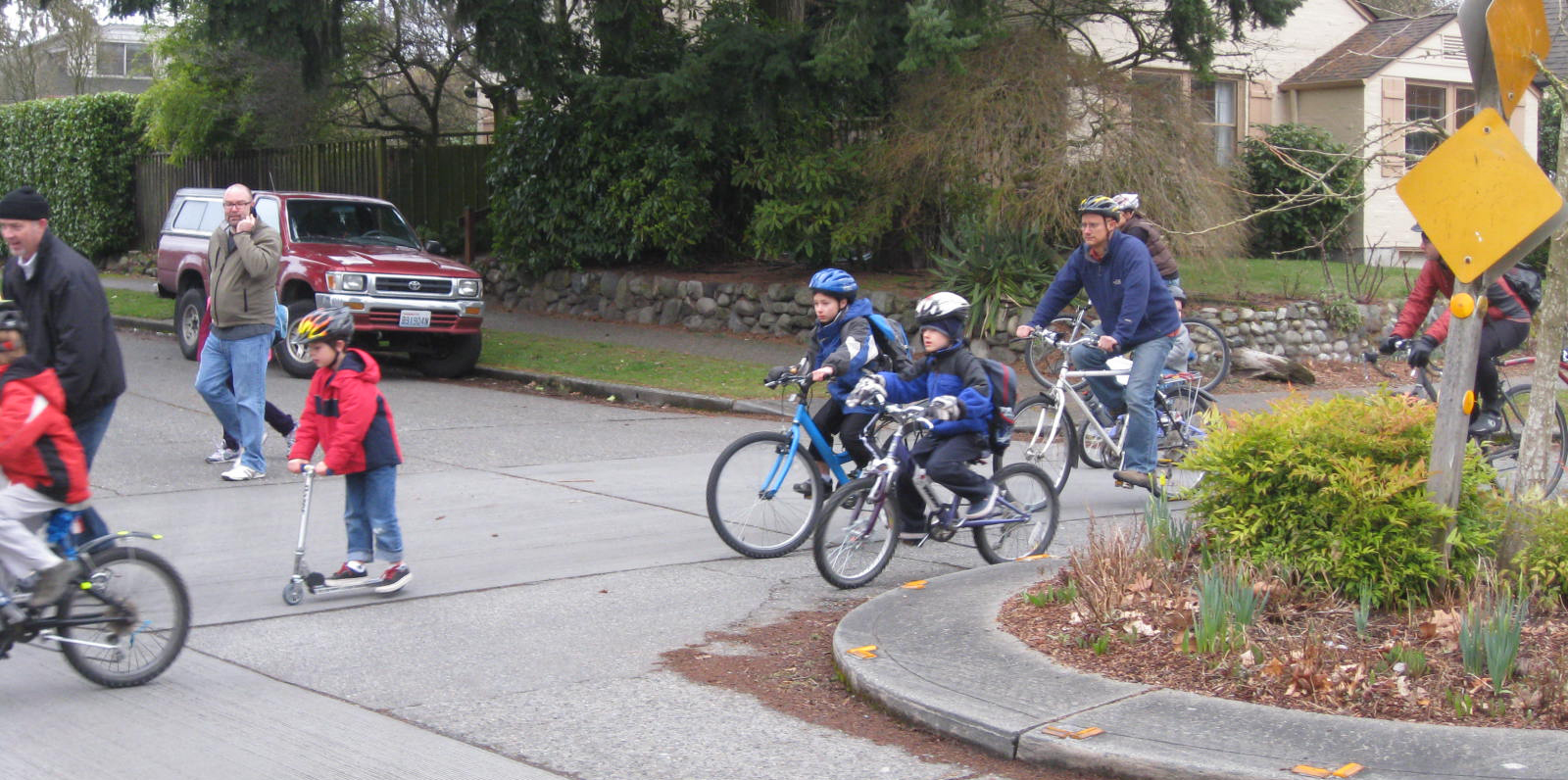 Children and adults ride their bikes around a roundabout in a suburban neighborhood.