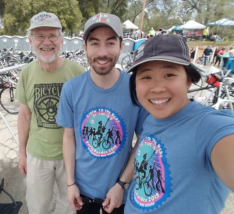 Three people - a young Asian woman, a young white man, and an older white man with bikes parked behind them.
