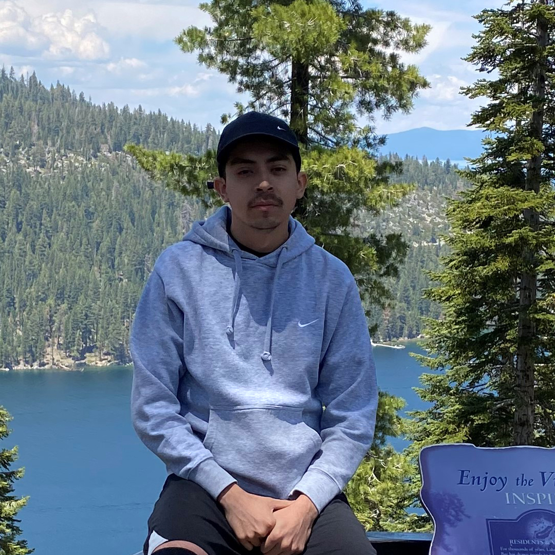 A young man sits with a lake and pine trees behind him.
