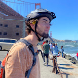 Man wearing a bike helmet stands by the bay with the Golden Gate Bridge in the background.