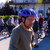 A man with a bike helmet stands with his bike. Other cyclists are in the background.