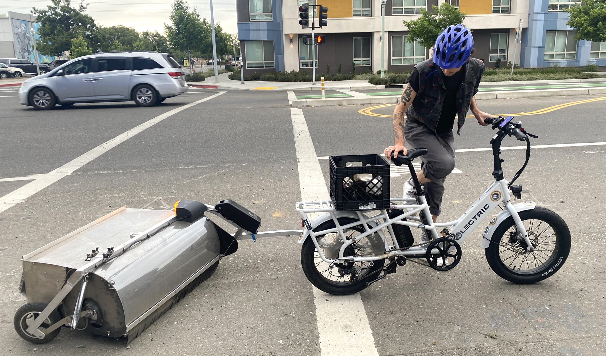 A man wearing a bike helmet and with tattooed arms sits on an ebike pulling a bike lane sweeper.