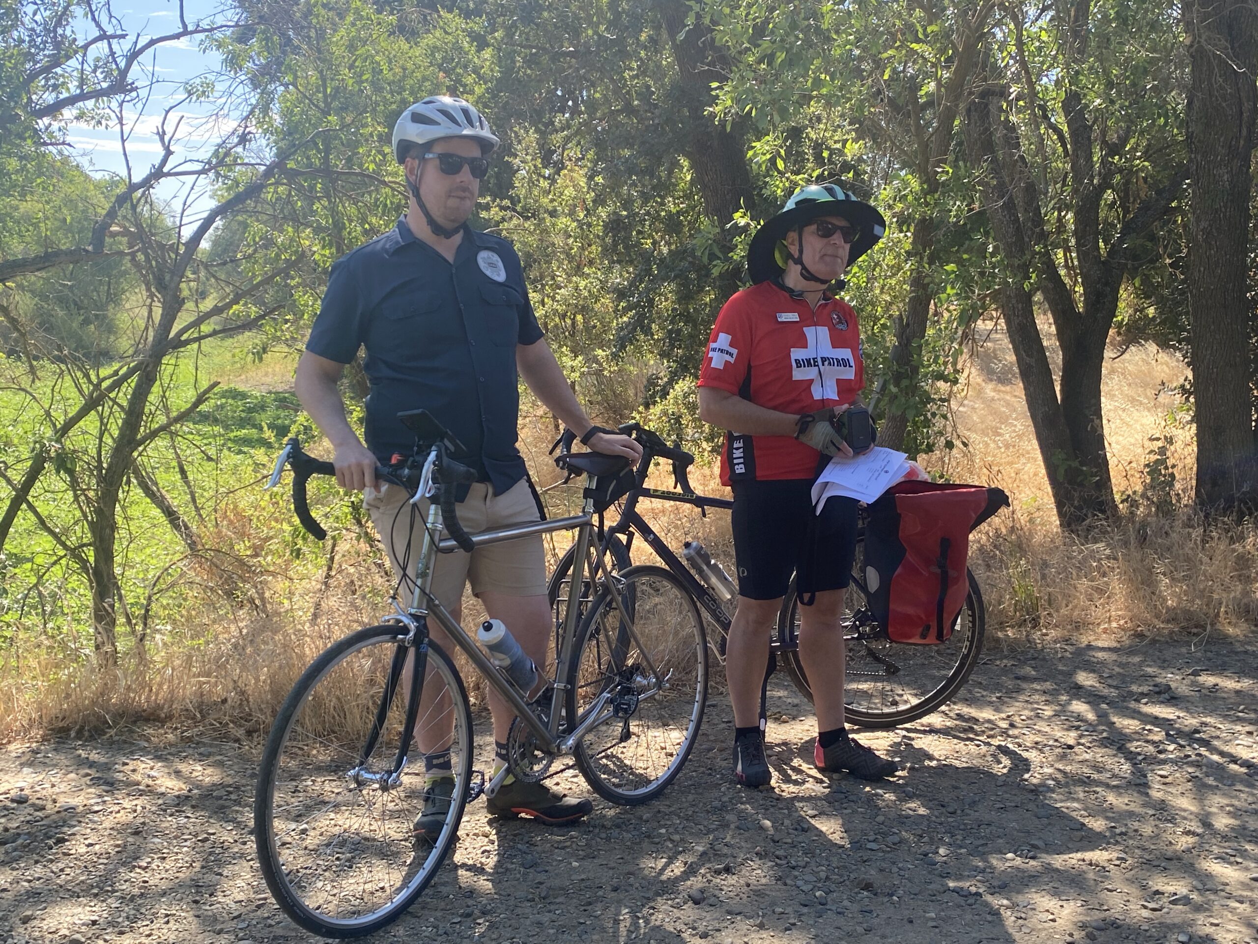 Two men stand by their bicycles on a dirt path in the woods.